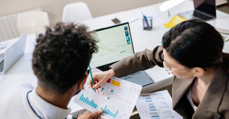 Two colleagues in a meeting room discussing financial charts and graphs on a laptop and paper.